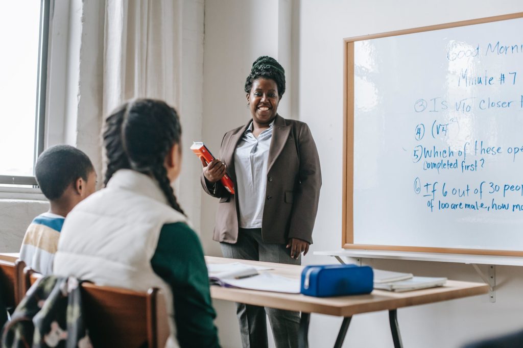 anonymous classmates sitting at table during math lesson with black female schoolteacher